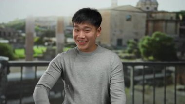 Gri kazaklı genç Çinli bir adam antik Roma harabelerinin önünde parmaklarıyla gülümsüyor ve işaret ediyor, güneşli bir arka planı var, kültürel ve tarihsel titreşimleri yakalıyor..