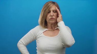 Orta yaşlı, çatık kaşlı beyaz kadın, parlak mavi stüdyo alanında çıplak elle tapınağa dokunuyor. Acı..