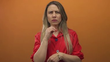 Woman covering her face with both hands in a photography studio with bright orange backdrop, wearing red blouse and tense posture; fear.