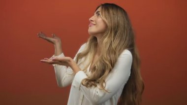 Beyaz bluzlu uzun saçlı kadın stüdyoda yukarı bakarken her iki avucunu havaya kaldırır; davetiye merakı açıklık sunumu.