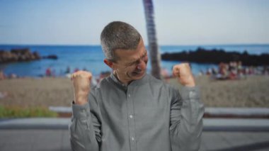 Sahilde sevinç ifade eden bir adam, yumruklarını deniz kenarına sıkıştırmış, açık havada mutluluğu yakalıyor, sakin deniz ve kumlu sahil berrak gökyüzünün altında görülebiliyor..