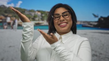 Gülümseyen bir kadın, güneşli bir sahilde okyanus ve tepelerin olduğu beyaz bir tişörtle mutlu bir sahil deneyimi sunuyor..