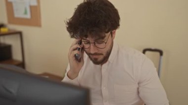 Ofis ortamında gözlüklü ve sakallı genç adam telefonda konuşuyor. Masa, bilgisayar ve raflarla çevrili, iş yerinde profesyonel iletişimle meşgul..