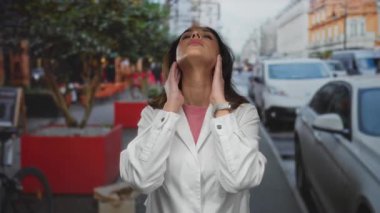 Şehir caddesinde beyaz laboratuvar önlüğü giyen kadın, şehir arabaları ve ağaçların arasında yoğun çevrenin altını çizerek hayal kırıklığı ve yorgunluk gösteriyor..