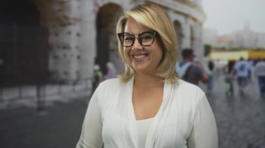 Gözlüklü sarışın kadın gündüz vakti kolezyum binasının dışındaki kamerayı işaret ediyor; özgüveni vurgula.