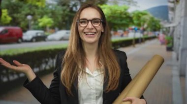 Gülümseyen bir kadın bir elinde karton tüp tutarken diğer elinde el hareketleriyle sokaklara el sallıyor..