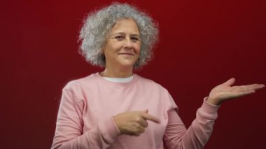 Gri saçlı bir kadın, kırmızı bir duvarın önünde, pembe giyinmiş, pozitif ifade ve güven gösteren, modern tarzda izole edilmiş, el hareketleriyle parmağını kaldırıyor..