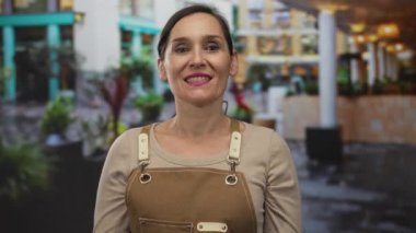 Şaşkın ifadeli önlüklü bir kadın şehir çevresi ve bitkilerle çevrili şehir caddesinde dikiliyor, açık havada canlı bir etkileşim sergiliyor..