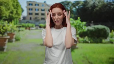 Başı ağrıyan beyaz gömlekli kadın, yeşil manzaralı, tarihi bina geçmişi olan güneşli bir parkta duruyor. Kentsel alandaki duyguları ve açık hava ortamını vurguluyor..