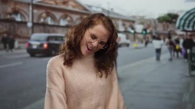 Gündüz gözlük ve süveter takan genç bir kadın şehir geçmişine sahip ve arkasında yürüyen bulanık yayalar.