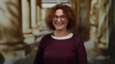 Kıvırcık saçlı, gülümseyen ve üniversite ortamında el kol hareketi yapan bir kadın akademik ortamda neşe saçıyor..