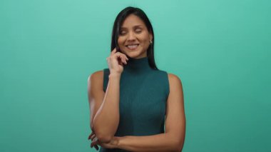 İzole edilmiş yeşil bir arka plana neşeli bir şekilde gülümseyen genç enerjisini vurgulayan düşünceli bir pozla parmağını yanağına koymuş bir kadın.