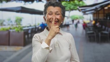Gri saçlı yaşlı kadın hareketli bir şehir caddesinde sessiz bir jest yapıyor. Şehir manzarası ve açık hava manzarası ile çevrili, pozitif enerji ve bilgelik yayıyor..