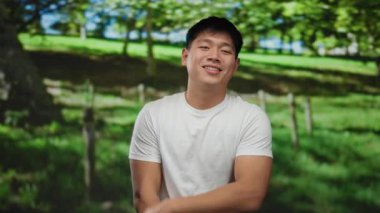 Açık havada güneşli bir günde yeşil bir parkta gülümseyen ve poz veren beyaz gömlekli genç bir adam..