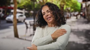 Beyaz kazaklı bir kadın, gözleri kapalı bir caddede kendini kucaklıyor, mutluluk, rahatlama, kıvırcık saçlar, olgun güzellik, açık hava, sakin bir yüz ifadesi, gündüz ayarları..