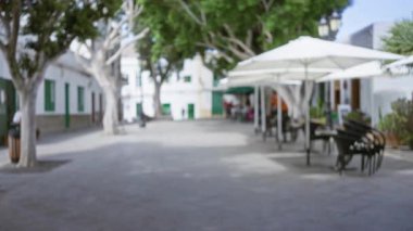 Lanzarote 'de bulanık bir açık hava sahnesi. Güneşli bir günde, küçük bir meydanda ağaçların, binaların, bankların ve şemsiyelerin altındaki masaların görüntüsü bulanık.