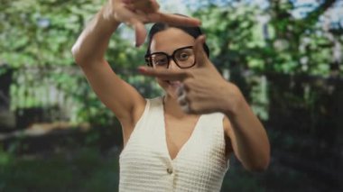 Güneşli bir günde yeşil arka planlı, parktaki genç bir kadın gözlük takıyor ve açık havada günlük giysiler giyiyor, gülümsüyor ve yaratıcılığa odaklanıyor, anın tadını çıkarıyor..