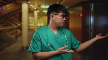 Yeşil önlüklü genç Çinli adam modern bir otelin içini işaret ederek, sağlık hizmetleri profesyonelliği ve sofistike otel tasarımının karışımını vurguluyor..
