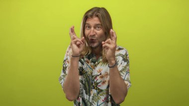 Çiçekli gömlek giyen uzun saçlı sarışın adam, parlak yeşil stüdyoda çıplak elleriyle çapraz parmaklar tutuyordu..