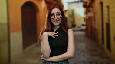 Kızıl saçlı ve gözlüklü bir kadın şehir caddesinde dikiliyor. Sıradan siyah bir bluzla düşünceli görünüyor. Gündüz huzurlu bir şehir atmosferini yansıtıyor..