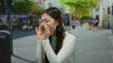 Şehirdeki şehir caddesinde bağıran bir kadın. Etrafı araçlar ve yeşilliklerle çevrili. Beyaz uzun kollu bluz giyiyor..