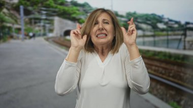 Beyaz gömlekli bir kadın endişeli bir ifadeyle parmaklarını caddede demiryolu raylarının üzerinde dışarıda, platformun yakınında duruyor. Umut..