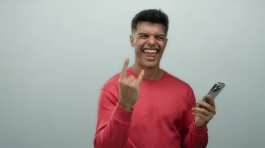 Kırmızı kazaklı genç İspanyol adam parmaklarıyla boynuz hareketi yaparken beyaz arka plana karşı akıllı telefon tutarak neşe ve heyecanı ifade ediyor..