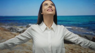 Sahilde güneşin tadını çıkaran bir kadın, açık kollarıyla mavi gökyüzünün altında beyaz gömlek giyiyor kayalık sahilde yaz tatili seyahati, dışarıda huzurlu sakin bir manzara.