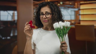 Sıcak bir restoranda, beyaz çiçekli ve yüzük kutulu bir kadın. Nişan hakkında şaşkınlık ve düşünce sergiliyor..