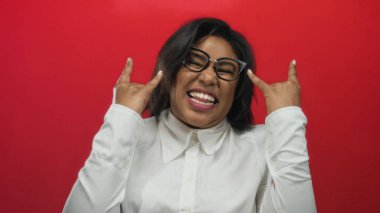 Beyaz tişörtlü, kırmızı arka planda genç bir Latin kadın elleriyle kaya jesti yapıyor, enerjik bir şekilde gülümsüyor, neşeli ve kendinden emin, iri yarı gözlüklü tarzını sergiliyor..