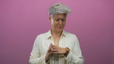 Kısa gri saçlı, beyaz tişörtlü bir kadın stüdyoda parlak pembe arka planlı bir burun çimdikliyor..