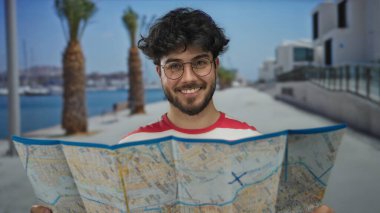 Sakallı genç adam güneşli caddede harita çalışıyor. Rıhtım ve modern binaların yanında palmiye ağaçları var..