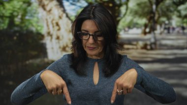 Gözlüklü ve uzun kollu mavi tişörtlü bir kadın yeşil parkta parmaklarını aşağı doğru işaret ediyor..