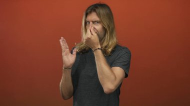 Sarışın adam kaşlarını çatar ve turuncu stüdyoda burnunu çimdikler. Gri gömlekli, uzun saçlı, omuzlarının üzerinde tiksinti vardır..