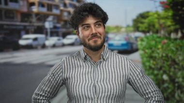 Sırıtkan, çizgili tişörtlü, sakallı genç İspanyol adam şehir manzarası bulanık arka planı ve yeşilliği olan hareketli bir sokakta güvenli bir şekilde dikiliyor..