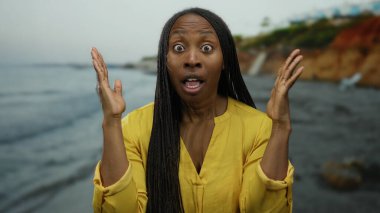 Sahilde dikilen, sarı tişörtlü, saçları örgülü kayalık sahil şeridine karşı duygularını ifade eden ve bulutlu gökyüzünün altında dalgalanan bir kadın..