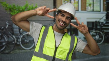 Beyaz kasketli ve yansıtıcı yelek giyen mühendis iki eliyle şehir sokaklarında barış işareti yapıyor; iyimserlik.