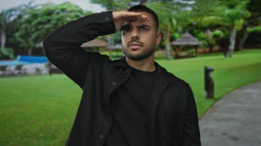 Ormandaki yeşil ağaçların arasındaki parlak güneş ışığına karşı gözlerini gölgeleyen adam parkı temizliyor..