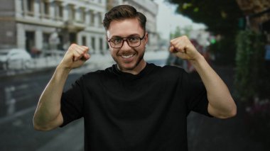 Sakallı ve gözlüklü genç adam bir şehir caddesinde başarıyı kutluyor, kendine güveni tam bir ifade sergiliyor..