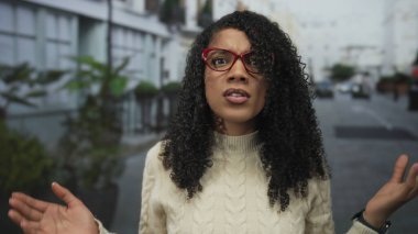 Kırmızı gözlüklü kadın, şehir caddesinde saksı bitkilerinin yanında iki elini kaldırır, açık avuç içi hareketi gösterir; şaşkınlık.