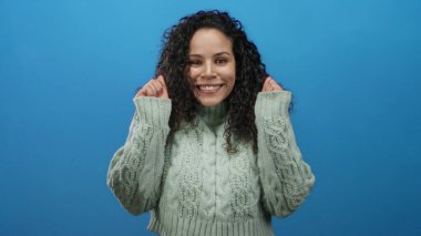 Canlı mavi arka planda, rahat süveter giyen bir kadın stüdyo ortamında izole edilmiş mutluluğu ifade ediyor.