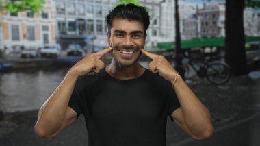 Genç adam yaygın bir şekilde gülümsüyor ve yoğun şehir caddesinde siyah tişört giyip, parmaklarını ağıza veriyor..