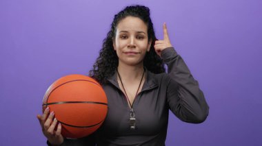 Mor bir duvarın önünde basketbol oynayan bir kadın şakağına işaret ederken, spor bağlamında düşünceli bir poz öneriyor, ıslıkla günlük spor kıyafetleri giyiyor..