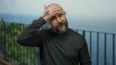 Beyaz sakallı bir adam, sıcak güneşin altında düşünceli bir şekilde, deniz kenarında geziniyor..