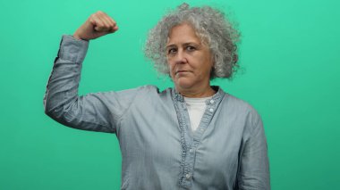 Gri saçlı yaşlı bir kadın, izole edilmiş yeşil arka plan duvarına karşı güçlü bir jest yapıyor..
