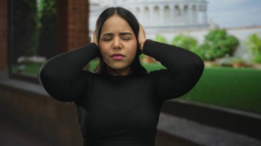 Latin kökenli genç bir kadın dışarıda kulaklarını kapatıyor. Kule 'nin sokak manzarası sıkıntılı görünüyor..