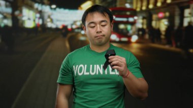 Yeşil tişörtlü genç gönüllü adam, arka planda bulanık ışıklar, farkındalığı ve hatıraları simgeleyen hareketli bir şehir caddesinde gece vakti siyah kurdele tutuyor..