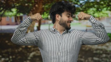 Sakallı, gülümseyen ve kaslarını kasan genç adam yeşil ağaçların altında çizgili bir gömlek giyerken..