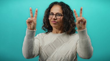 Kıvırcık saçlı kadın zafer işareti yaparken canlı mavi bir arka plana gülümsüyor, kendine güveni ve pozitifliği var..