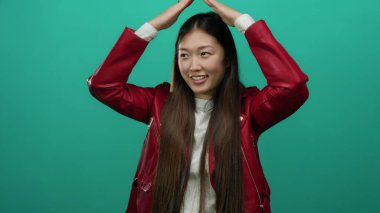 Kırmızı ceket giymiş, yeşil arka planda elleri şekilli poz veren bir kadın.